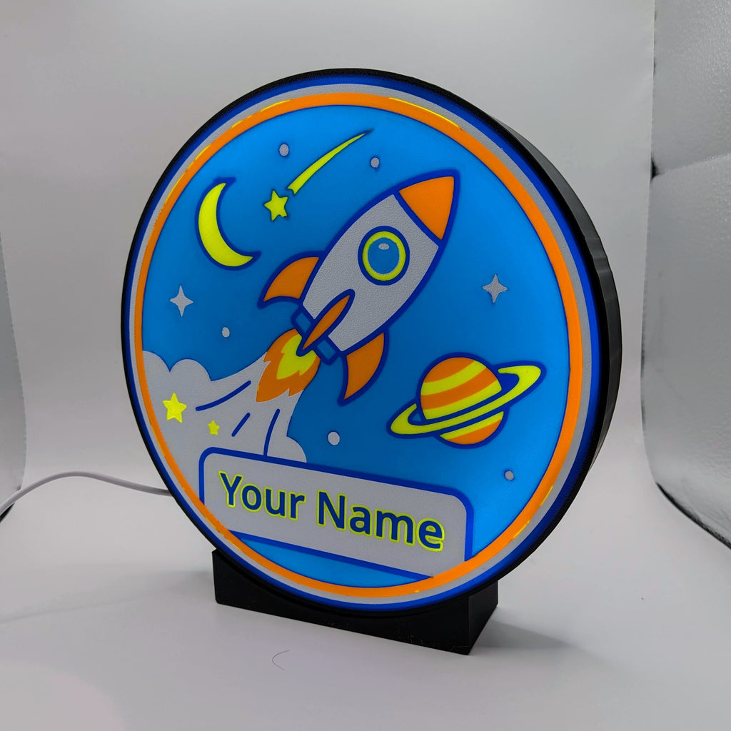 Lampada LED rotonda blu con razzo, pianeta saturno e scritta 'Your Name' su supporto