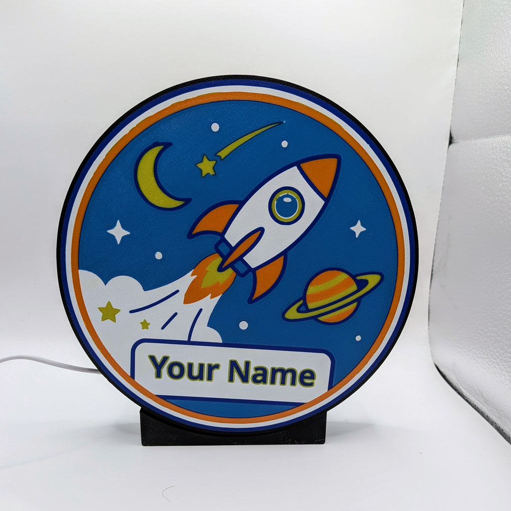 Lampada LED rotonda blu con razzo e cometa, scritta 'Your Name' e base nera
