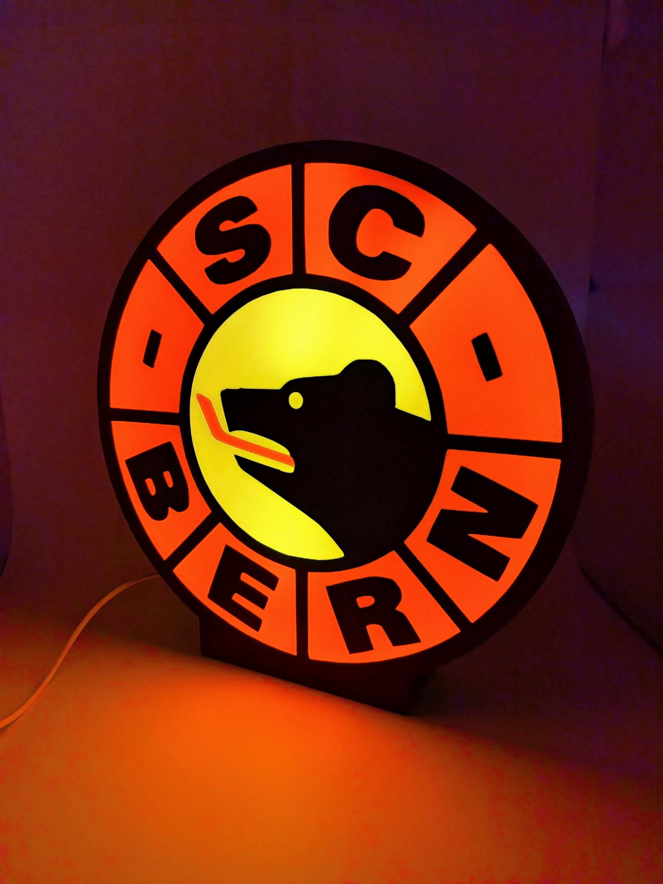 Lampada LED rotonda SC Bern con bordo arancione, cerchio giallo e supporto nero.
